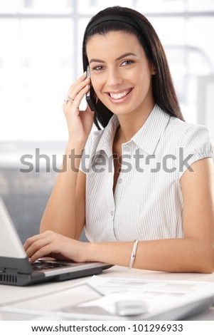 Cheerful office assistant using laptop computer on mobile phone call, laughing happily at camera.