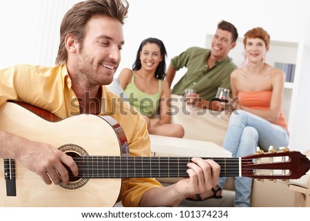 Young happy friends having party, one playing guitar, the others listening.