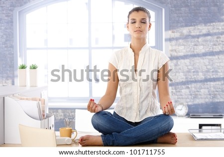 Pretty girl sitting on top of desk in bright office, meditating.