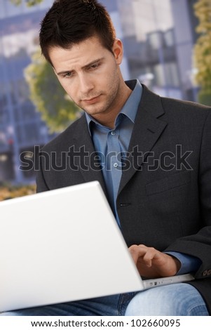 Businessman sitting outside of office, working on laptop computer, concentrating.