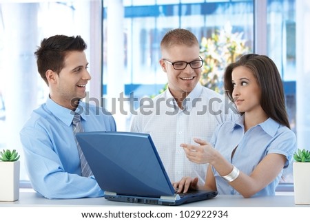 Happy businesspeople standing together, talking at work, using laptop computer, laughing, pointing at screen.