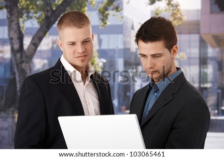 Two businessmen standing with laptop computer outside of office, looking at screen, smiling.