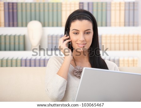 Smiling woman sitting at home looking at laptop computer screen, using mobile phone.