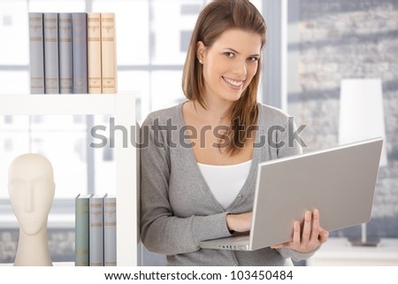 Happy woman standing at bookcase with laptop computer handheld, smiling at camera.