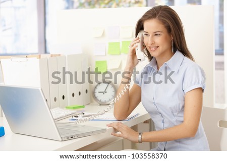 Attractive businesswoman on landline phone call, sitting at desk, looking at screen.