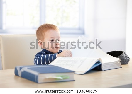 Little kid playing schoolboy, sitting at table, looking at encyclopedia.