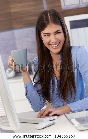 Happy businesswoman sitting at office desk, working, holding coffee mug, laughing.