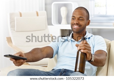 Smiling afro man enjoying leisure time in living room, with beer and remote control.