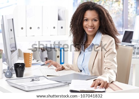 Portrait of beautiful smiling afro-american office worker sitting at desk, using computer.