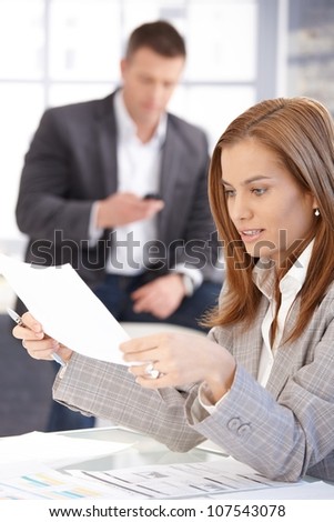 Young female working with papers in office, man texting in background.