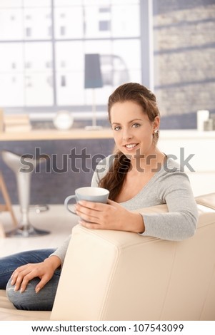 Attractive woman sitting on sofa at home, drinking tea, smiling.