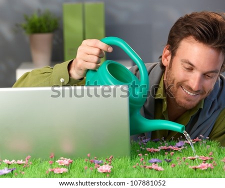 Cheerful man watering spring field table, smiling, laptop computer and pink flowers.
