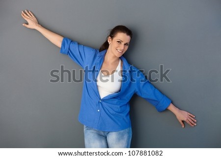 Portrait of attractive mid-adult woman posing at grey wall with arms wide open, smiling at camera.