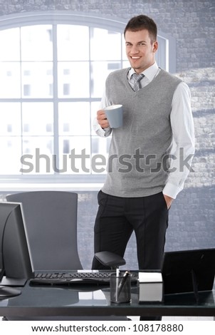 Casual young businessman standing in bright office, drinking tea, smiling.