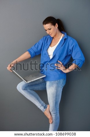 Pretty mid-adult woman standing with laptop computer, casual pose, looking at screen, smiling.