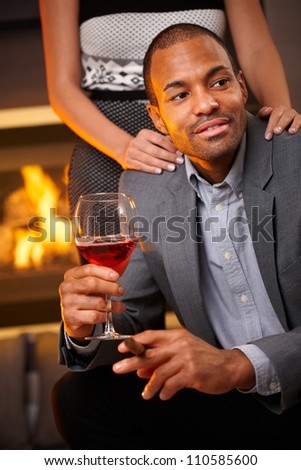 Goodlooking ethnic man sitting by fireplace smoking cigar, drinking wine, girlfriend standing behind.