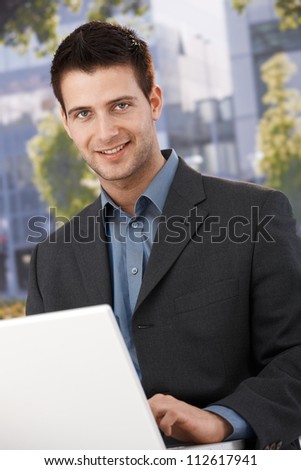 Smiling businessman sitting outside of office building, using laptop computer, looking at camera.