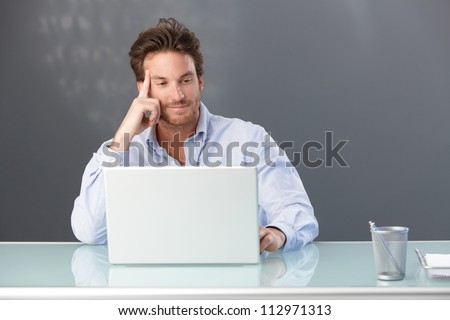 Smiling handsome male office worker sitting at desk looking at laptop computer screen.
