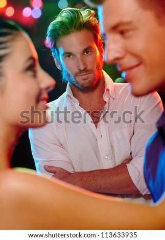 Jealous man looking at dancing couple, flirting girlfriend in nightclub.
