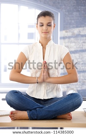 Young attractive female sitting on top of desk, meditating.