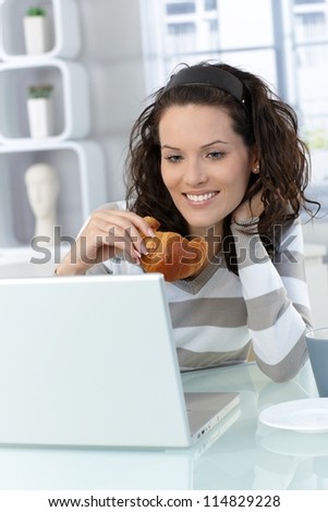 Happy woman eating croissant at home, using laptop computer, smiling.