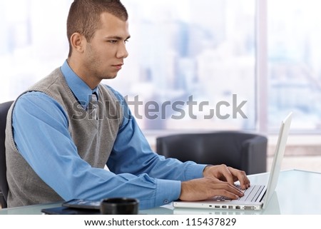 Young businessman concentrating on working with laptop computer in skyscraper office.