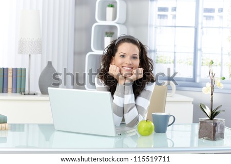 Portrait of happy young woman sitting in bright study, laptop computer, mug, green apple on desk.