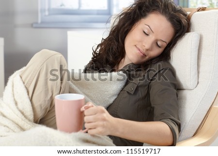 Young woman relaxing in armchair under blanket at home, holding tea mug, smiling, daydreaming.