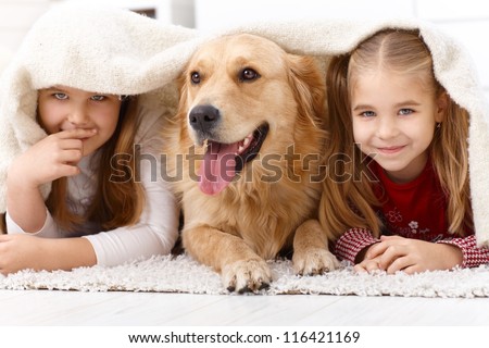 Cute Little Girls Having Fun With Golden Retriever, Lying Prone On Floor At Home Under Blanket, Smiling.