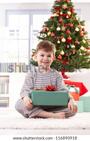Portrait of happy little kid sitting in pyjama in christmas morning holding gift parcel, smiling at camera.