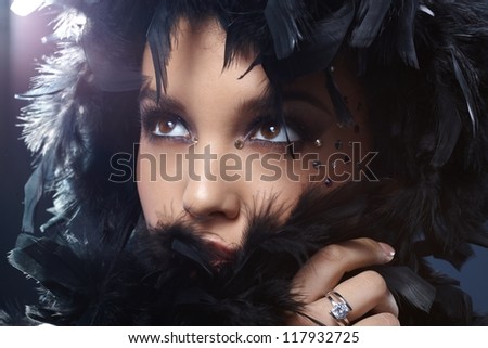 Beauty in elegant makeup with rhinestones hiding in feather boa, looking up.
