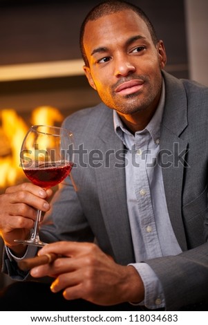 Handsome black man sitting by fireplace smoking cigar, drinking wine.