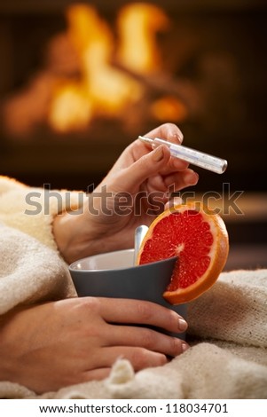 Checking temperature, illness, winter cold, female hands holding tea mug with blood orange and thermometer in front of fireplace.