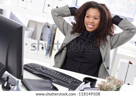 Beautiful afro businesswoman sitting at desk in office, smiling, stretching.