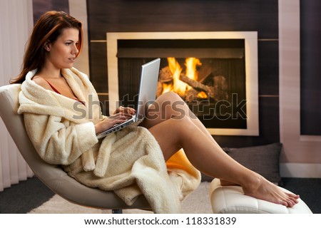 Hot woman sitting in bathrobe using laptop computer in front of fireplace, enjoying winter leisure.