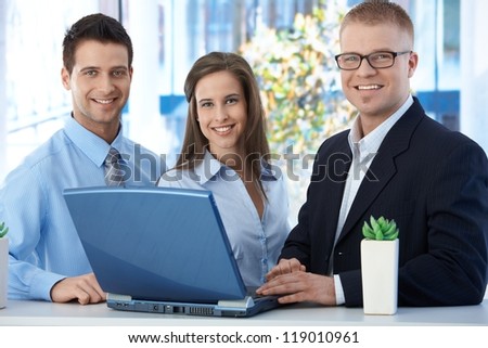 Portrait of young businessteam standing in office, using laptop computer, smiling at camera.