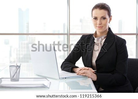 Portrait of elegant businesswoman sitting at desk with laptop computer, smiling at camera, copyspace.