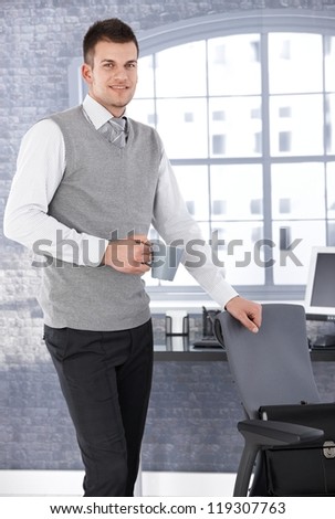 Young businessman standing in bright office, drinking tea, smiling.