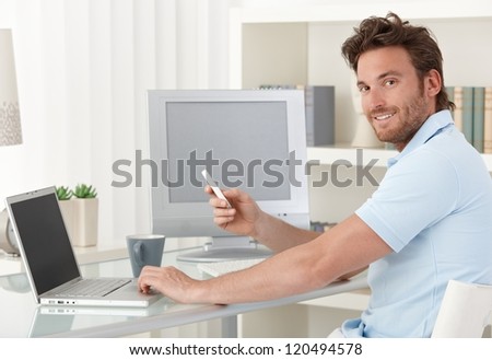 Man sitting at desk using laptop computer and phone at home, smiling at camera. Blank space on screens for your logo or image.