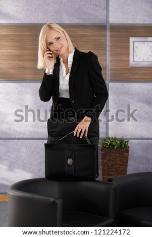 Businesswoman standing in office lobby, on mobile phone call, opening briefcase, taking out personal organizer, smiling.