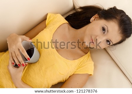 Young female resting on sofa, holding coffee cup, looking at camera, view from above.