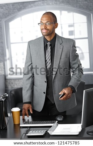 Elegant businessman standing at office desk with mobile phone handheld, briefcase and coffee on table.