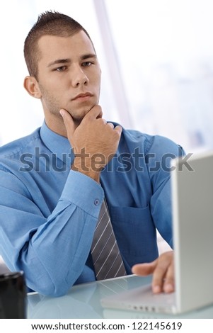 Serious young businessman at desk with laptop computer, thinking.