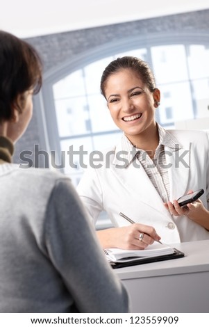 Pretty receptionist taking notes at counter, holding mobile phone, smiling at client.