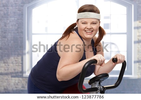 Happy fat woman training on exercise bike, smiling.