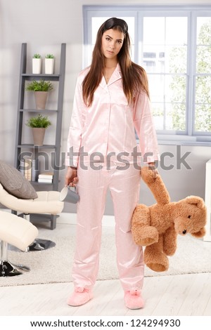 Portrait of attractive sleepy woman standing in pyjama with teddy bear and coffee mug handheld in living room.