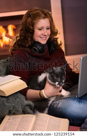 Smiling teenage girl sitting at fireplace at home learning with laptop and books. Happy brainy preoccupation.