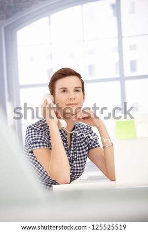 Assistant girl in focus sitting in bright office, listening to landline phone call, smiling.