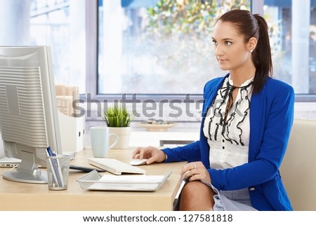 Pretty office worker at desk wearing fancy romantic shirt and cardigan, working on desktop computer.