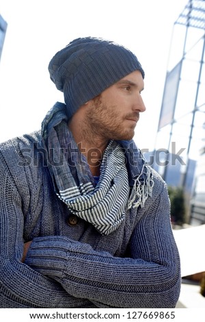 Outdoor portrait of trendy guy wearing scarf and hat, arms crossed.